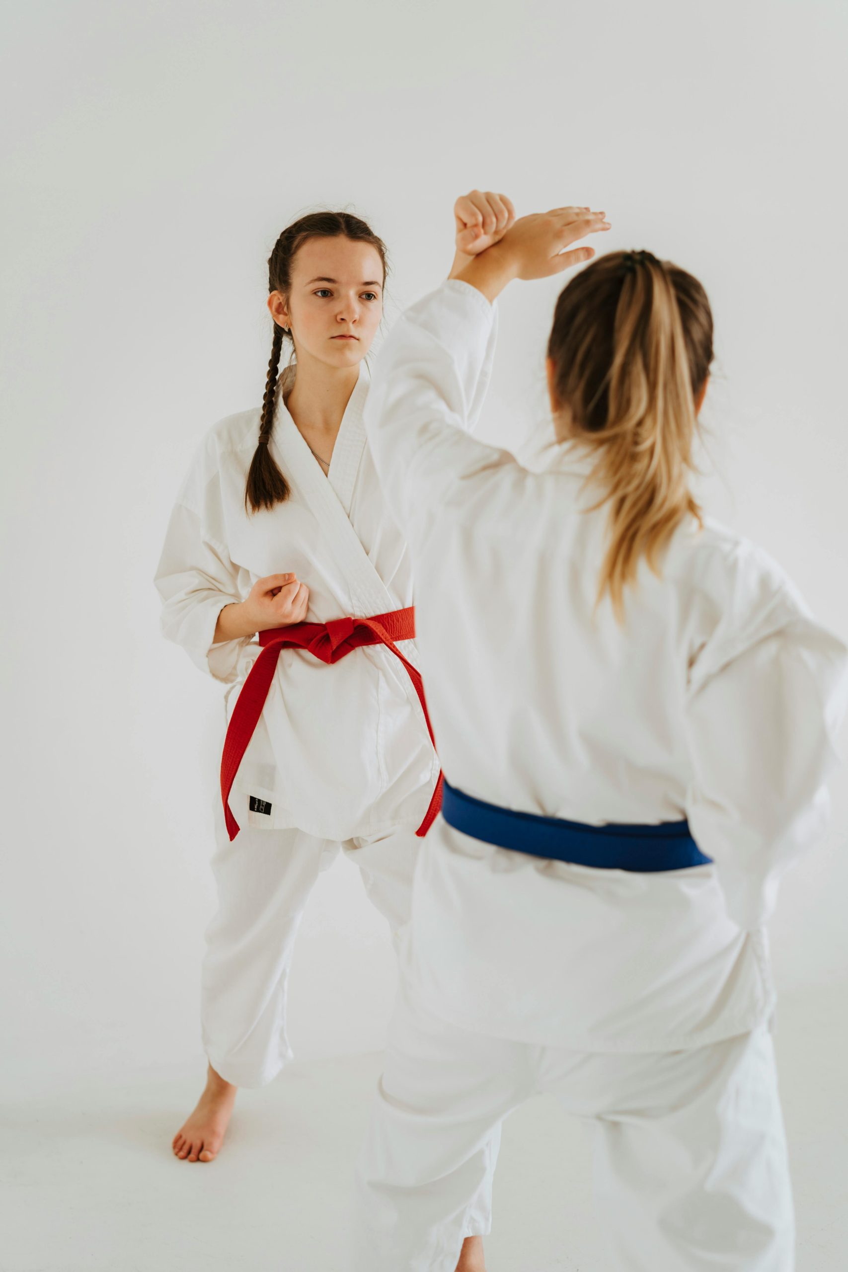 Two young women sparring in karate uniforms with red and blue belts on a white background.